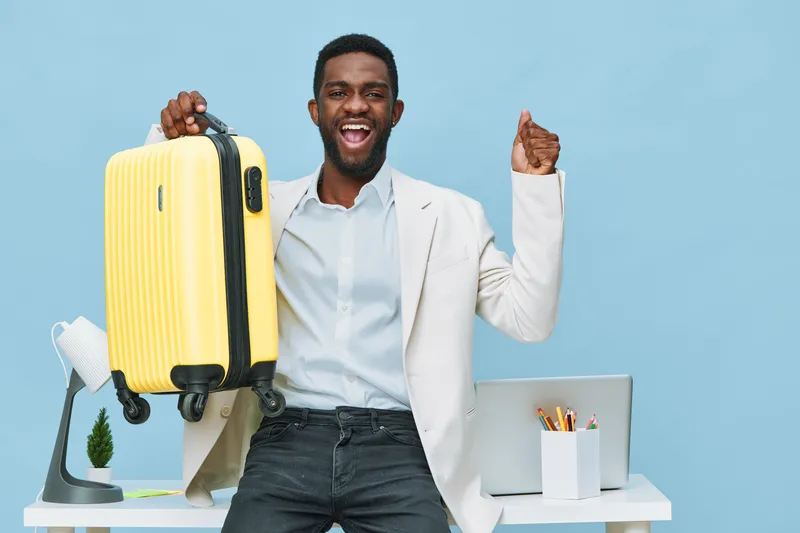 Smiling happy young man with dark skin holding yellow suitcase in modern office studio setting against blue background, wearing casual white shirt and blazer, expressing excitement and travel readines