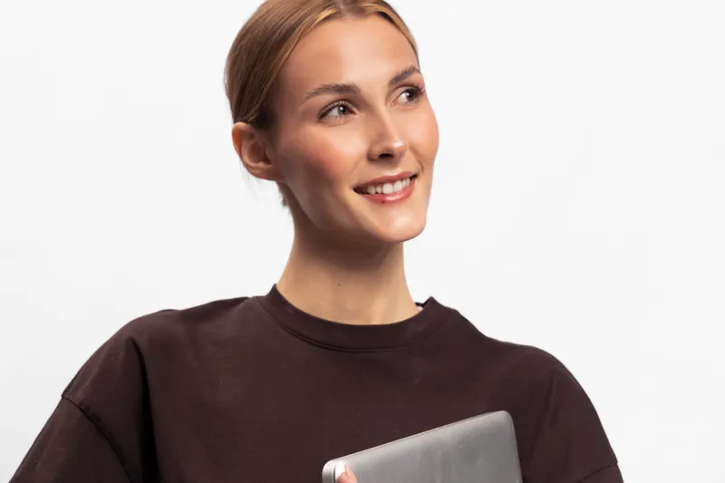 Smiling young woman with light brown hair holds silver laptop, looking aside. Studio portrait on white background. Concept for technology, business, education, and success.