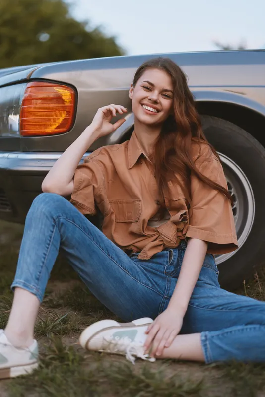 Young woman smiling in casual outfit sitting near vintage car surrounded by nature, conveying happiness, fashion, and summer vibes.