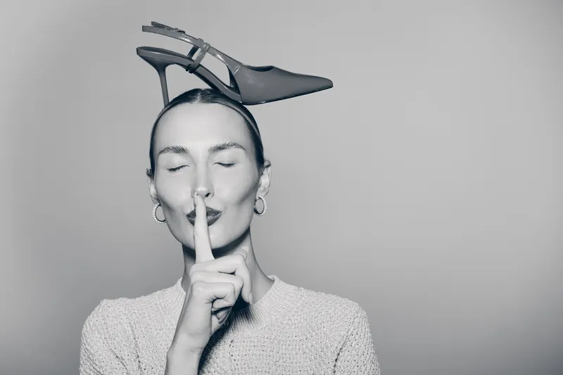 Young white woman with pale skin and closed eyes wears high heel shoe on head, finger on lips in silence gesture.