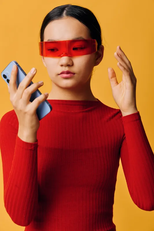 Woman with red sunglasses in red sweater holding smartphone and looking at screen against yellow background for technology, communication and fashion concept.
