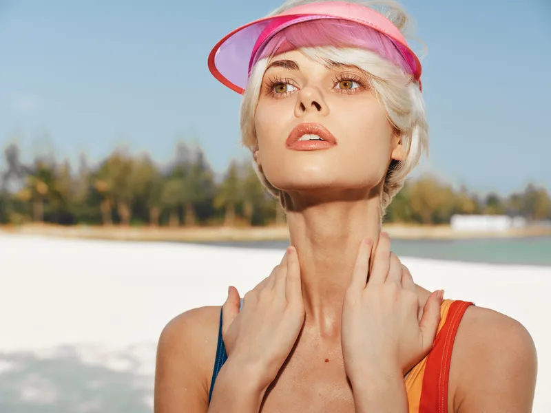 Portrait of a sunlit woman on a bright beach, wearing a pink visor and colorful swimsuit straps, gazing upward with a calm, confident expression under clear sky.