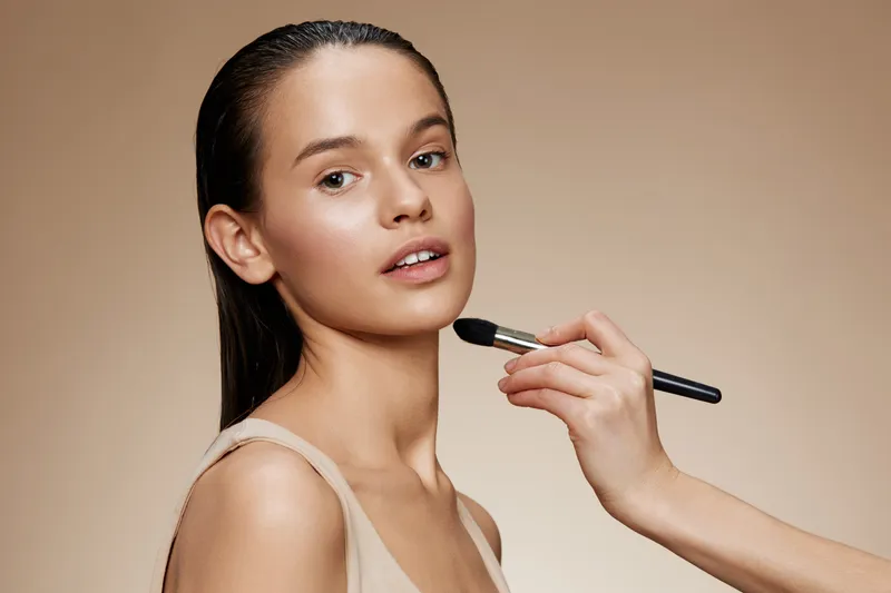 Beautiful female model receiving makeup application with natural tones on a clean background, conveying a fresh and polished look for beauty concepts