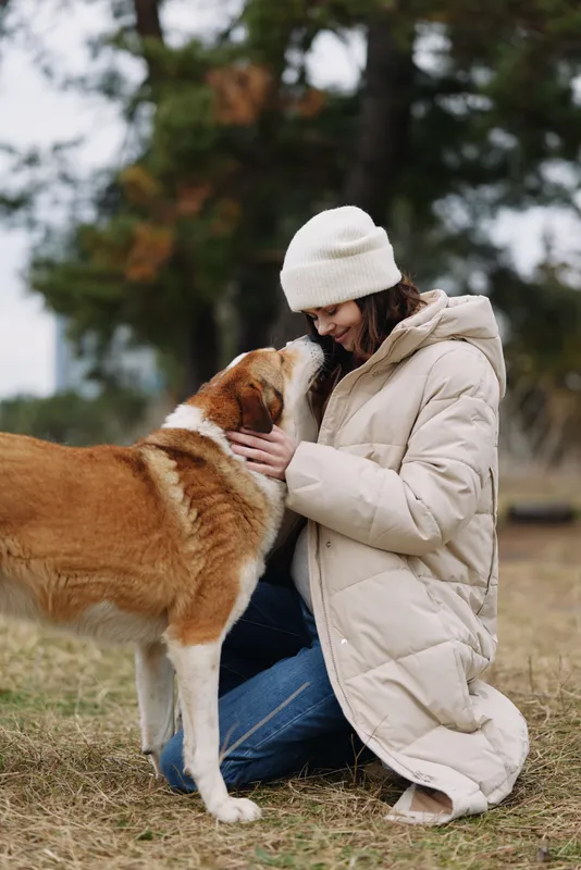 Dog and woman share affection outdoors as she kneels in a warm coat, offering gentle care on a chilly day. A serene, intimate moment highlighting companionship and natural bond.