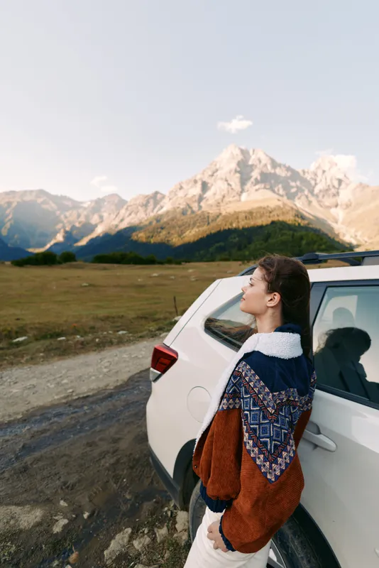 woman by car and mountains during travel in nature landscape, standing outdoors in cozy sweater. Scenic countryside portrait with meadow, peak horizon and relaxed autumn mood.