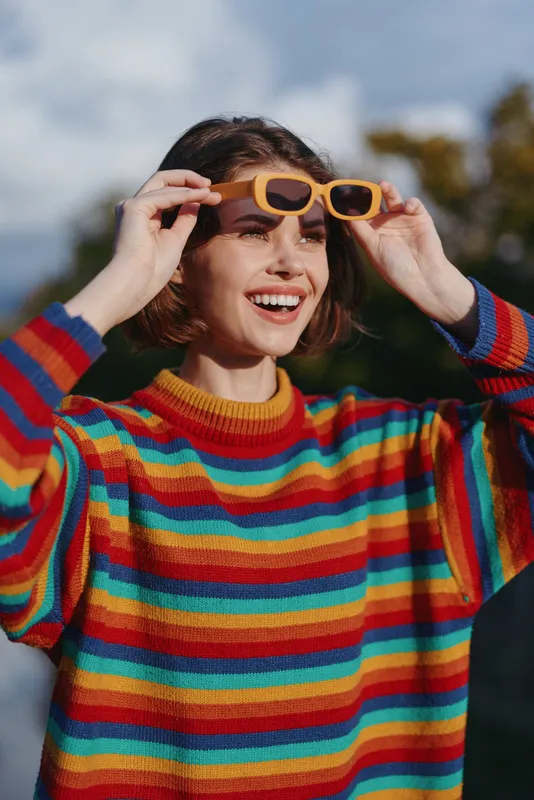 Young woman, smiling and joyful in her mid 20s wearing a colorful striped sweater and yellow sunglasses outdoors, casual outfit, playful portrait for lifestyle and fashion.