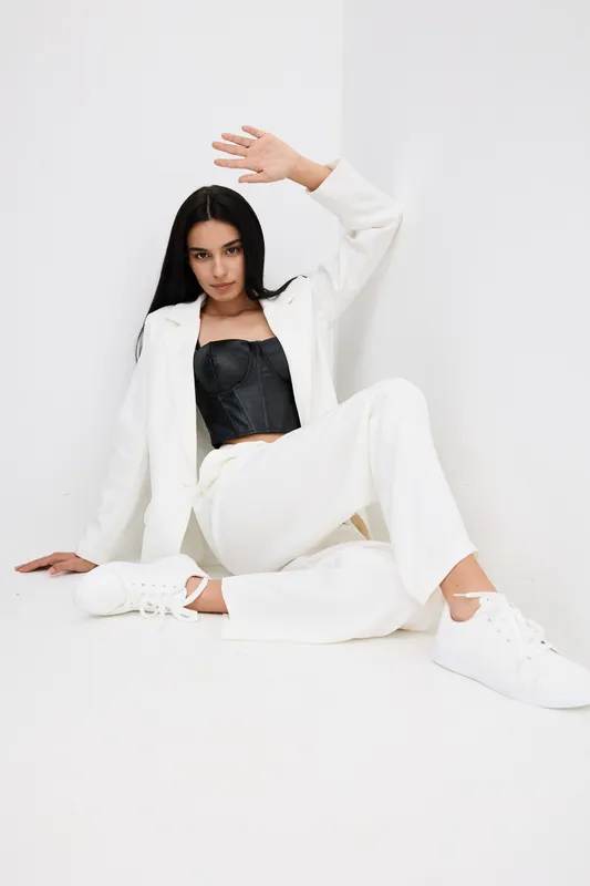 Young woman in white suit and black corset posing on white floor with casual white sneakers, minimalist background, stylish outfit, modern fashion look, relaxed mood.