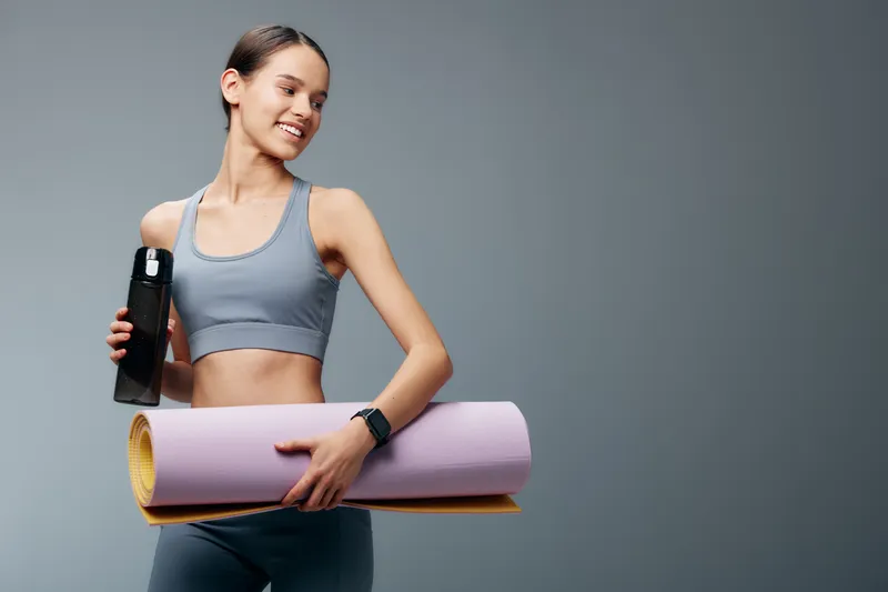 Fitness woman in athletic wear holding a water bottle and yoga mat, ready for workout, showcasing energy, focus and determination in a minimal studio setting.
