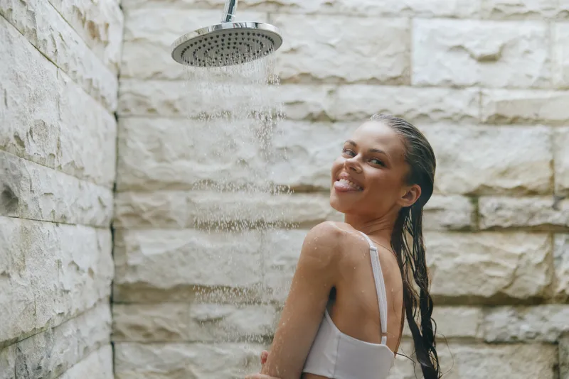 smiling woman enjoying outdoor shower under rainfall with natural stone wall background in casual swimwear, relaxed mood, bright natural light