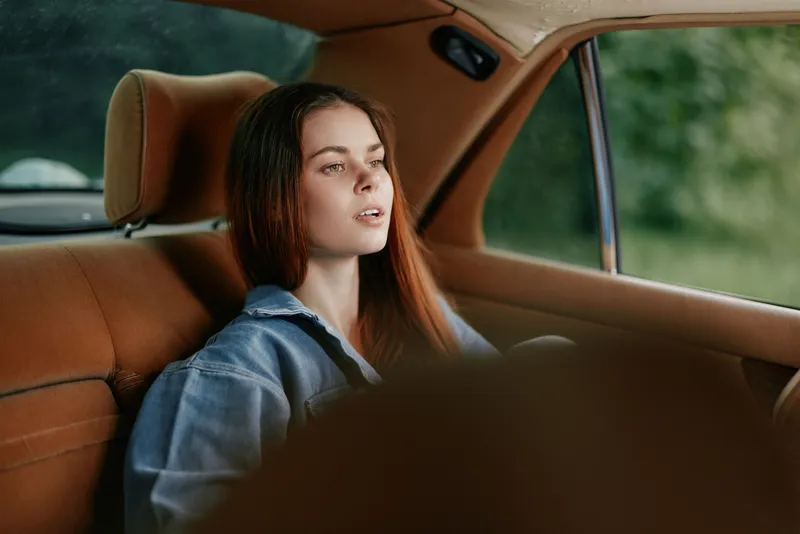 A serene young woman with long brown hair sitting in a vintage car, wearing a denim jacket, reflecting calmness and nostalgia amidst a lush green background.