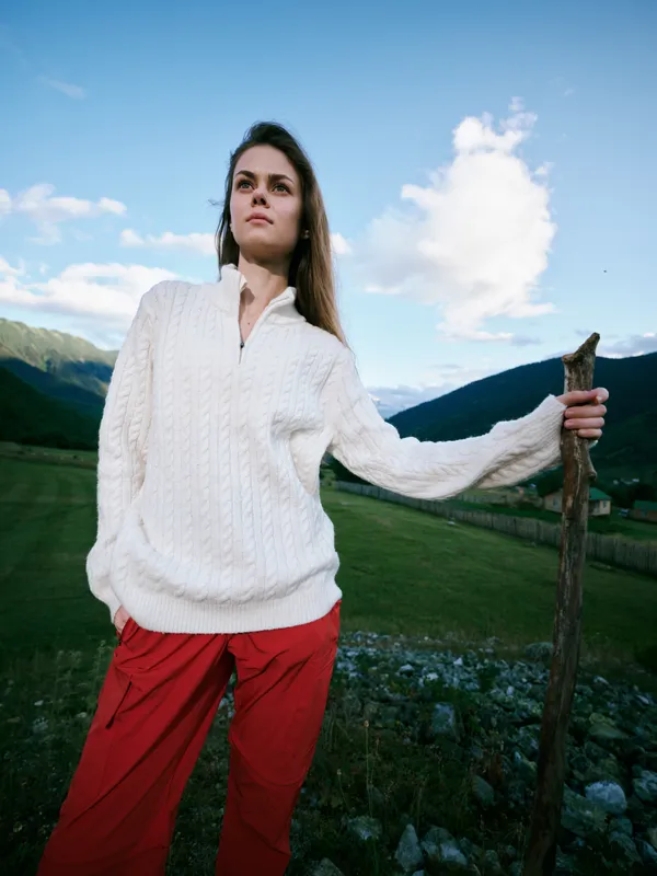 Woman in a white knit sweater and red pants stands in a grassy field with mountains in the background, holding a wooden staff under a blue sky, showcasing outdoor rustic landscape