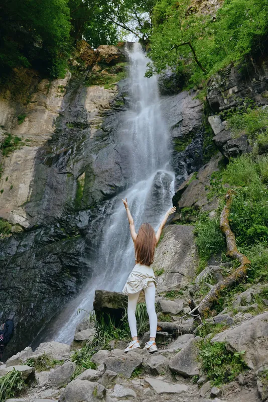 woman standing near tall waterfall with raised arms surrounded by lush green forest and rocky terrain during daytime