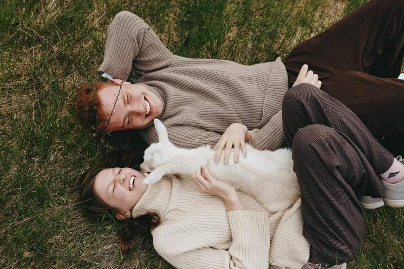 Two friends relax on the grass, smiling as they cradle a white cat between them, wearing cozy sweaters, casual mood, natural light, outdoor leisure, isolated on a park lawn with soft shadows and a