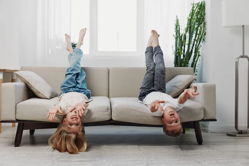 Two happy children play upside down on a beige sofa in a bright living room with casual clothes and joyful expressions. Concept of childhood fun and home leisure activity.