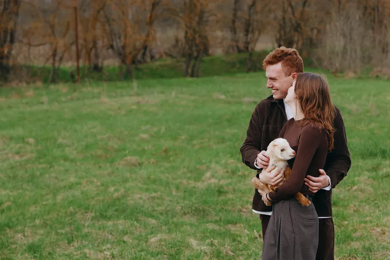 Young couple embraces in a green field while holding a small white baby goat, sharing a tender moment with natural surroundings and casual autumn clothing.