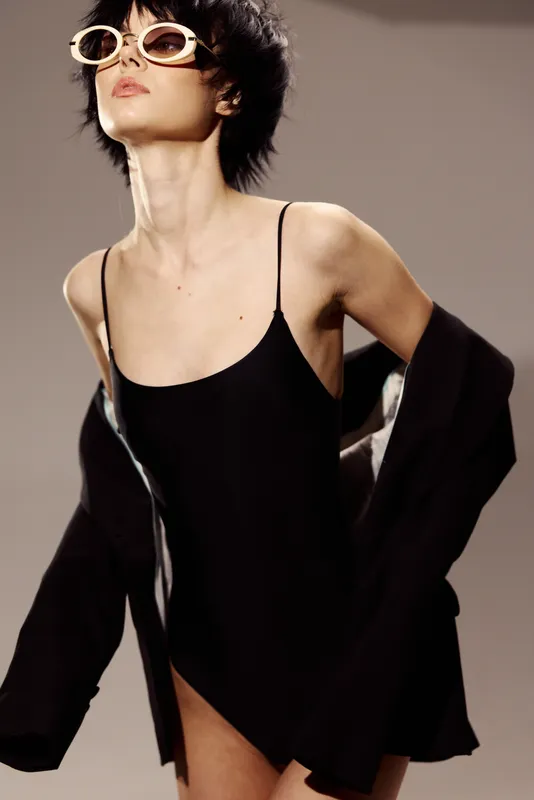 Stylish woman wearing black outfit and sunglasses, posing confidently indoors with neutral background and soft lighting.