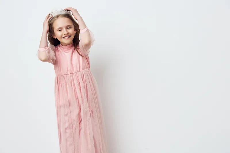 Cheerful girl in a pink dress wearing a crown, celebrating joy and innocence in a bright, minimalist setting Ideal for children s fashion and birthday themes