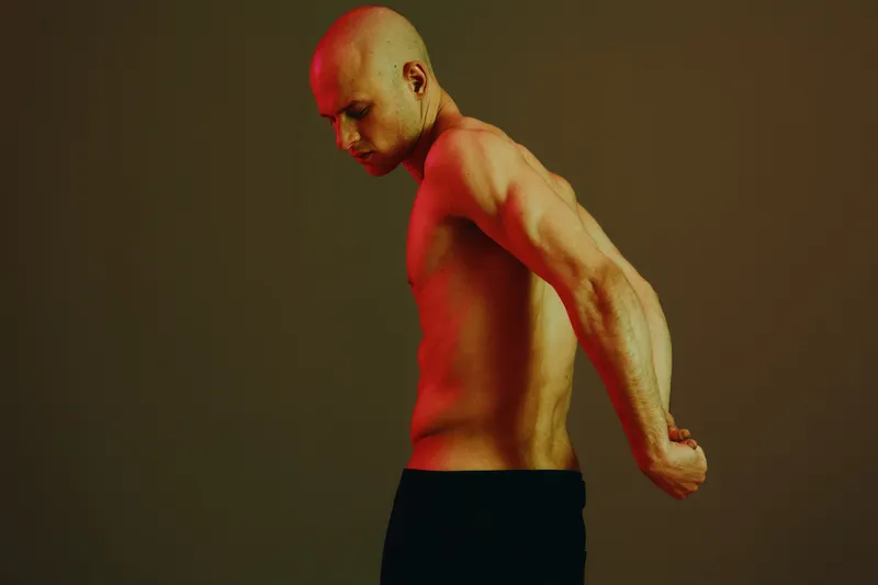 A shirtless man with a strong physique poses against a gradient background, showcasing strength and confidence. The striking red light emphasizes his muscular form.