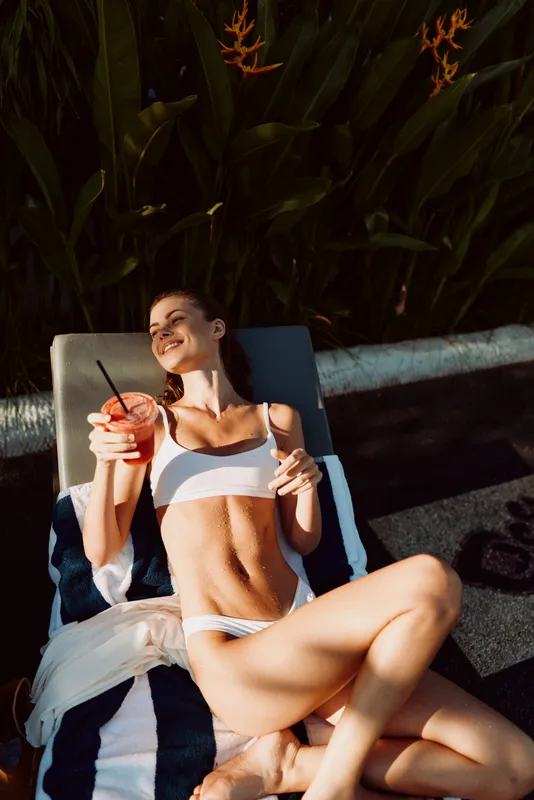 Poolside relaxation with a woman in a white bikini lounging on a chair, sipping a drink, enjoying warm sun, casual summer mood, towel nearby, stylish outdoor leisure moment.