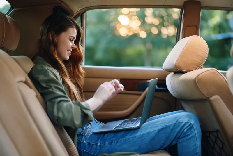 Young woman using laptop in a car's back seat during sunset, dressed casually for comfort and productivity, showcasing a relaxed and focused mood.