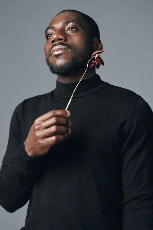 African man with dark skin holding dried flower in hand wearing black turtleneck sweater in creative conceptual visual style studio portrait with neutral background and thoughtful expression.