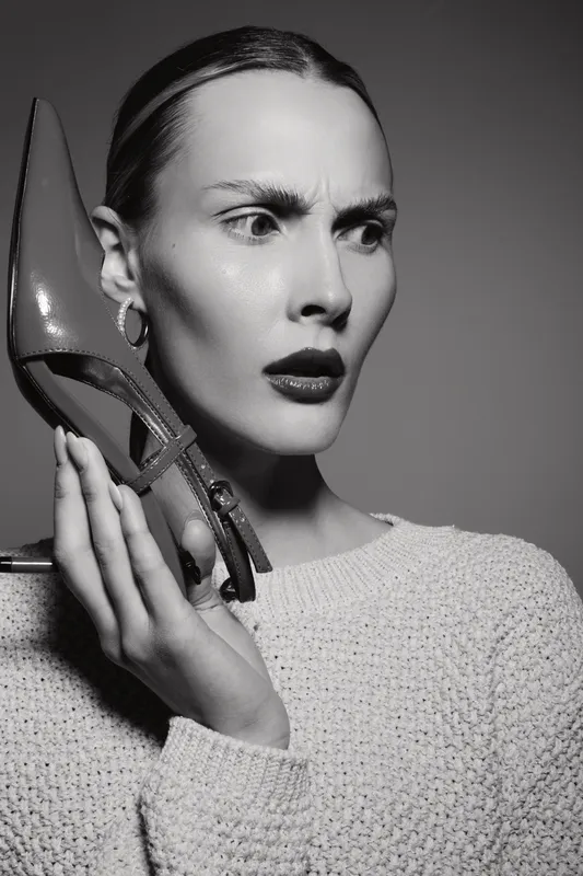 Young Caucasian woman with pale skin holds red shoe near ear with confused expression. Black and white portrait with serious mood and detailed facial features in studio lighting.