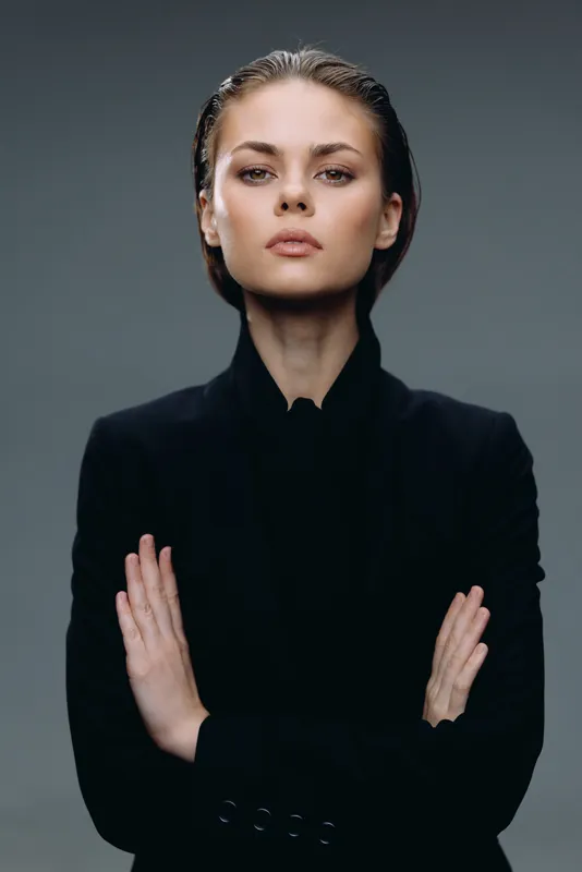 confident woman in black coat posing with crossed arms against gray background