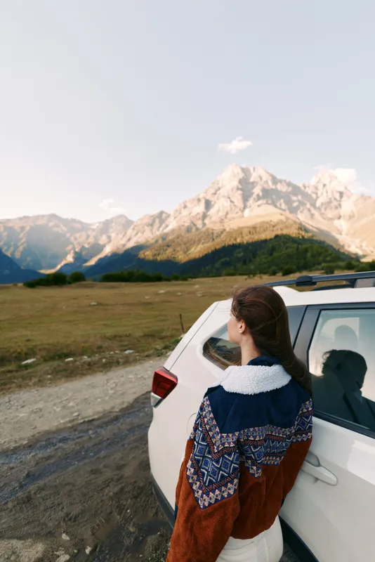 Woman in sweater beside car overlooking mountains and expansive landscape on a road trip, travel adventure and scenic journey outdoors at sunset with calm horizon and nature view.