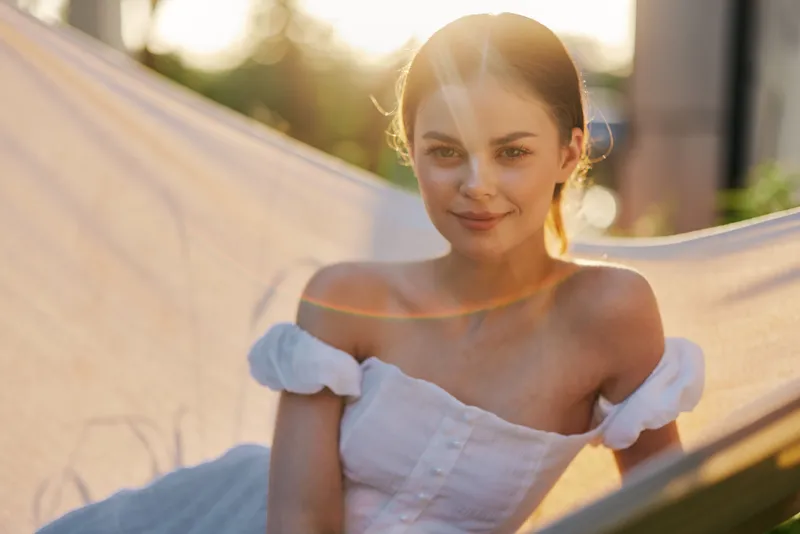Young woman in a white dress sitting in a hammock, enjoying a sunny day, evoking feelings of tranquility, joy, and warmth in a natural setting