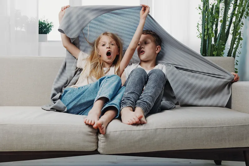 Two playful children sit barefoot on a beige sofa, covered with a gray blanket. The girl with blonde hair yawns while the boy beside her looks relaxed in a bright living room setting.