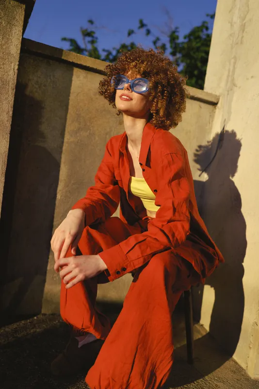 Candid woman outdoors in a vibrant orange suit sits on a chair, sunlit moment, relaxed pose, stylish sunglasses, warm shadows, urban backdrop, confident lifestyle