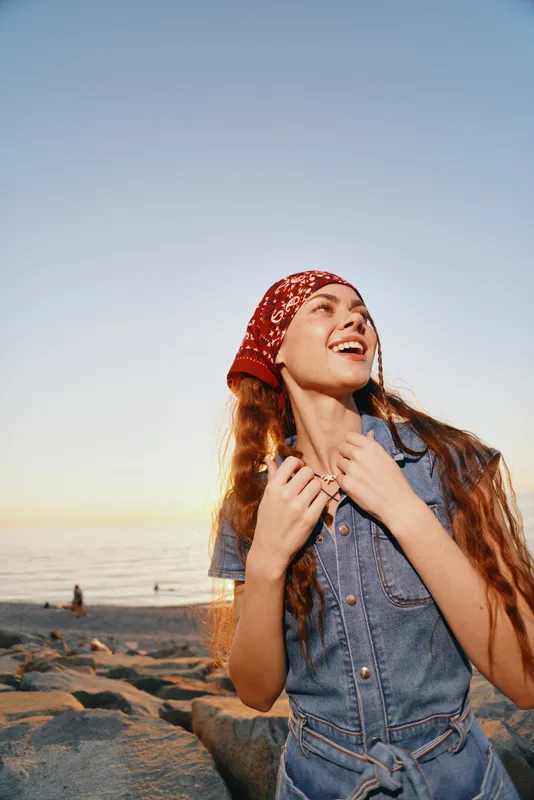 smiling woman with red bandana enjoying sunset at the beach wearing denim dress casual summer outdoors relaxation
