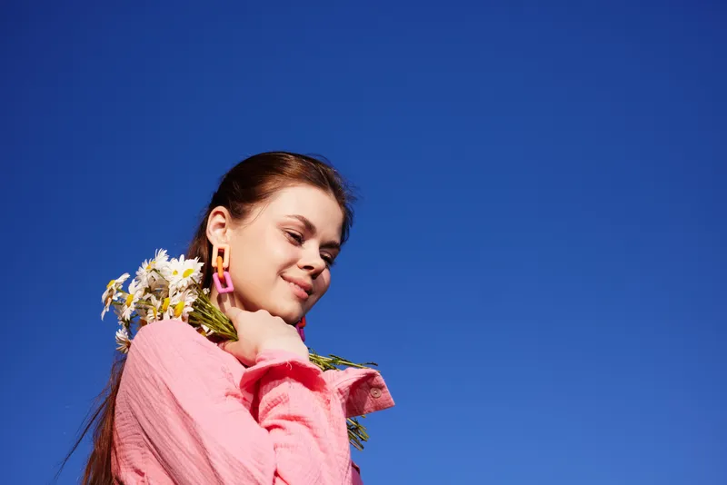 Young woman with flowers under a clear blue sky, wearing a pink blouse and smiling, conveying joy and freshness, ideal for seasonal and lifestyle themes