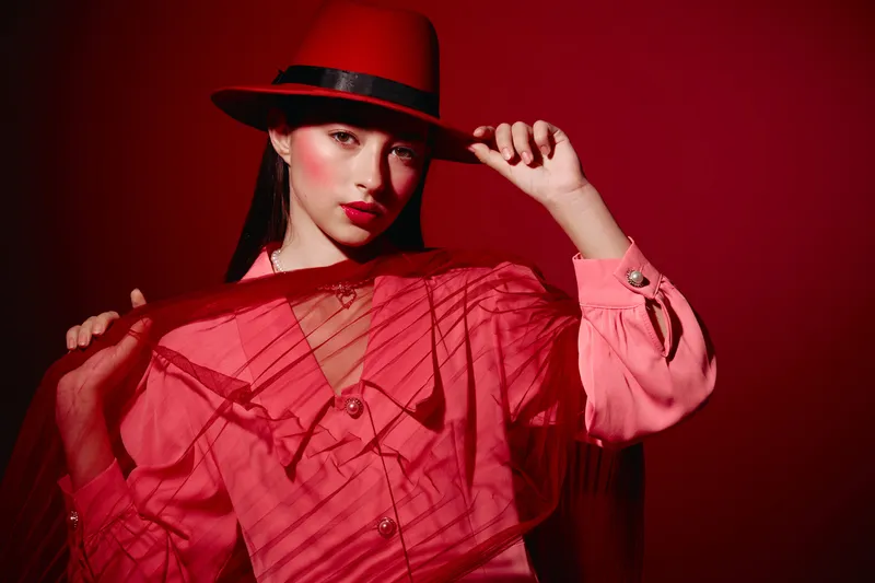 Young woman in stylish red hat and mesh outfit poses confidently against red background. Bold beauty and fashion creative emphasizing vibrant mood and color. Studio shot.
