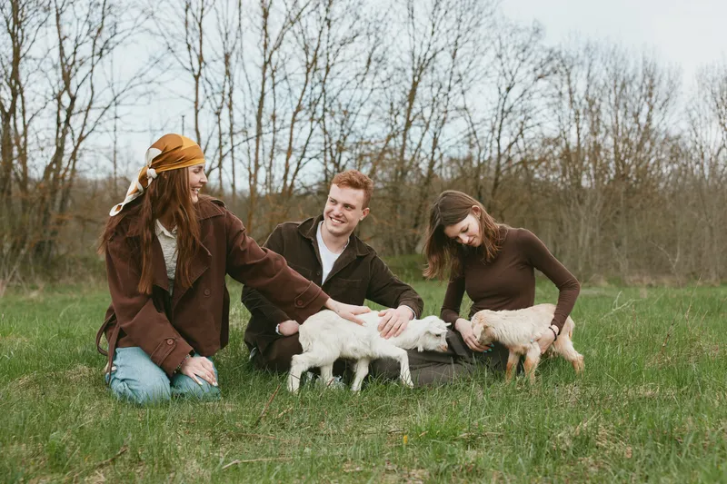 A joyful group of people chats and pets goats in a sunny pasture, capturing a rustic lifestyle moment and animal interaction in a casual farm setting that feels warm and welcoming.