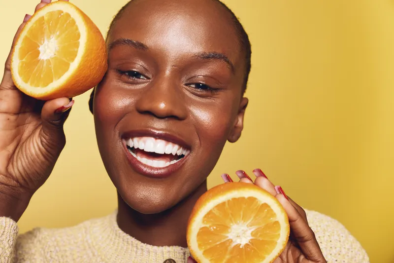 Happy African woman with dark skin holds two fresh orange halves near face, smiling with white teeth. Yellow background creates bright, cheerful concept of health, fruit, and wellness.
