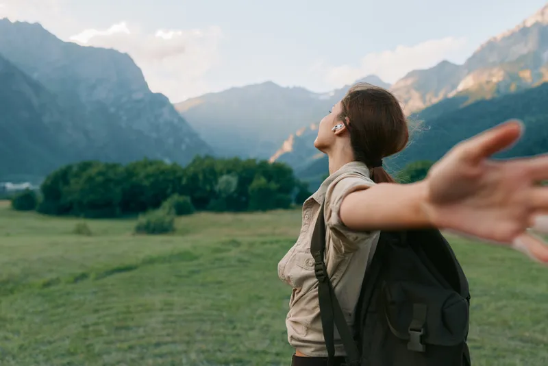 Young woman with backpack stands in a sunlit meadow with distant mountains, arms outstretched as if embracing a moment of carefree outdoor freedom.