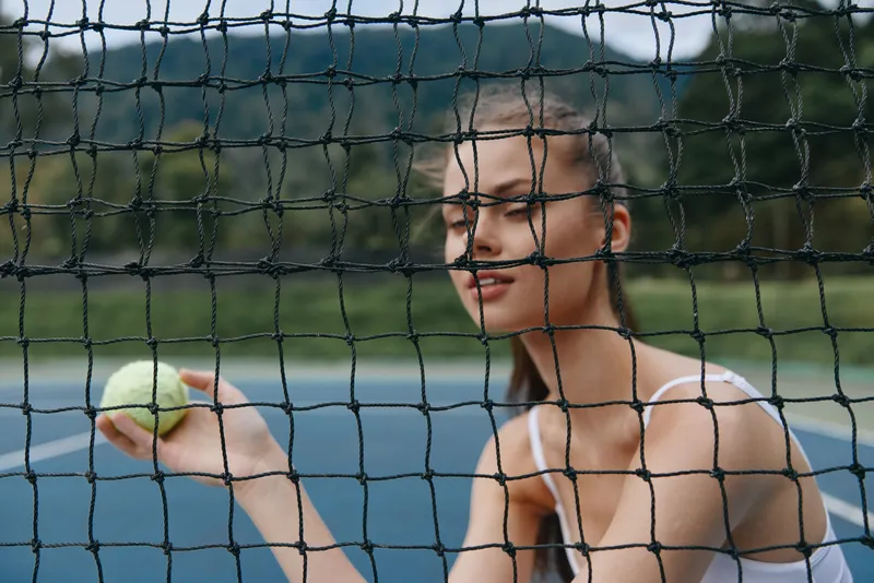 Young female tennis player smiling and holding a tennis ball behind the net on a sunny day, showcasing confidence and athleticism in a vibrant sports setting