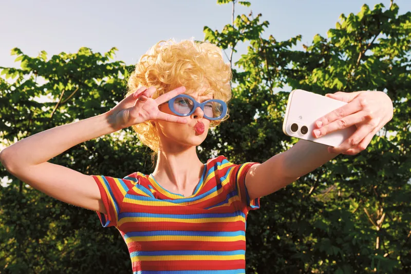 Selfie of a woman with curly blonde hair wearing a rainbow striped shirt and blue goggles outdoors, on a sunny day, vibrant greenery and playful mood, retro tone.