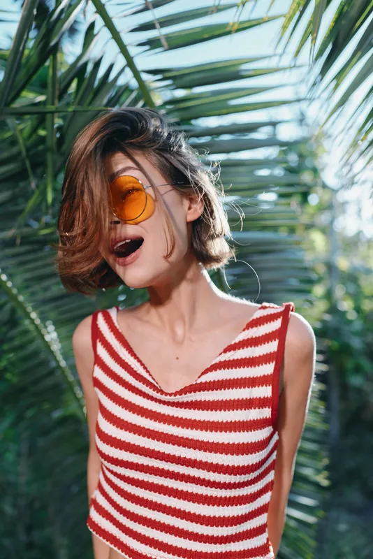 A stylish woman wearing a red and white striped tank top and orange sunglasses stands outdoors among lush tropical foliage, exuding a carefree summer vibe and confidence.
