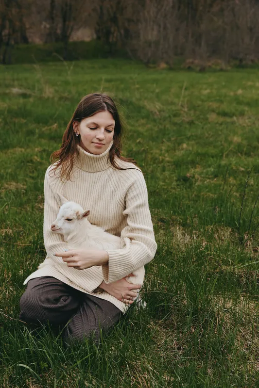 Bright outdoor scene with a woman in a cozy cream sweater sitting in a grassy field, gently holding a white goat kid, capturing a peaceful, tender lifestyle moment in sunlight