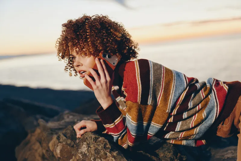 Woman lifestyle on a sunny beach, curly hair and striped sweater, holding a smartphone to her ear, relaxed during phone call outdoor, soft warm light, in calm mood, near quiet waves