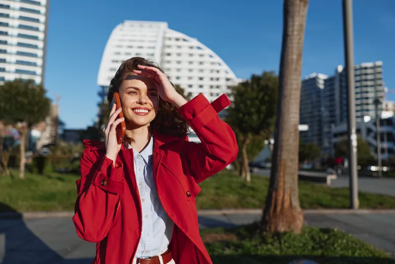 A young woman in a red trench coat stands in a sunny city park, shielding her eyes with one hand and talking on a phone, creating a confident, cheerful urban portrait.