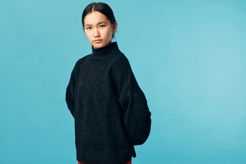 Young woman wearing black turtleneck sweater standing against blue background with serious expression. Portrait of female model with straight black hair and minimal makeup.