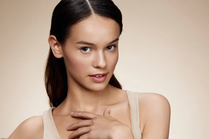 Beautiful young woman with natural makeup and a relaxed expression, showcasing healthy skin and subtle elegance against a soft beige background
