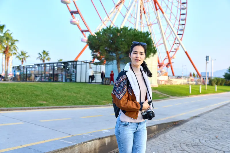 Woman with a camera stands outdoors near a ferris wheel at an amusement park, wearing a patterned jacket and jeans, capturing candid travel moments and sunny park ambience.