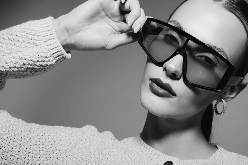 Close-up of a young Caucasian woman with fair skin wearing oversized sunglasses and a knitted sweater. Studio portrait with elegant makeup and fashion accessories on grey background.