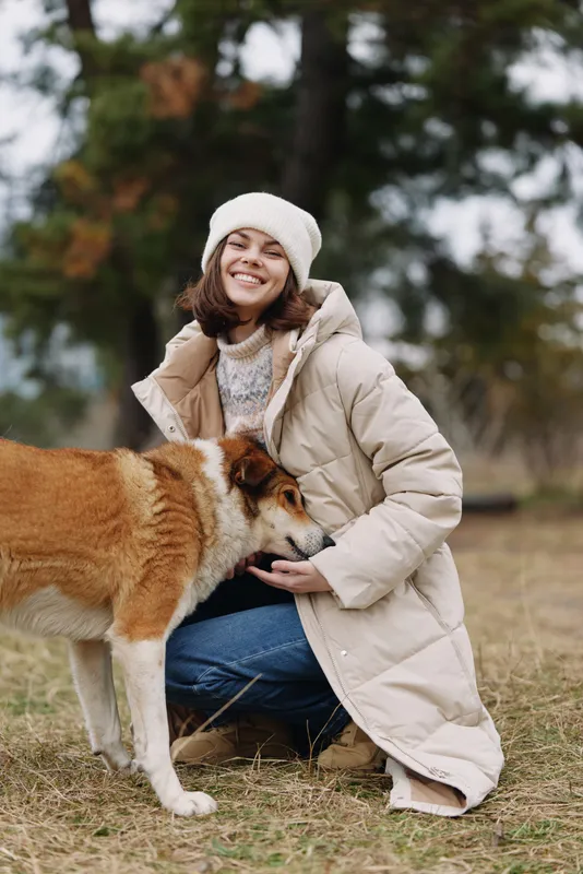 Woman kneeling in a park, wearing a beige quilted coat and knit hat, smiles while petting a friendly dog on a chilly day, conveying warmth and joyful bond.