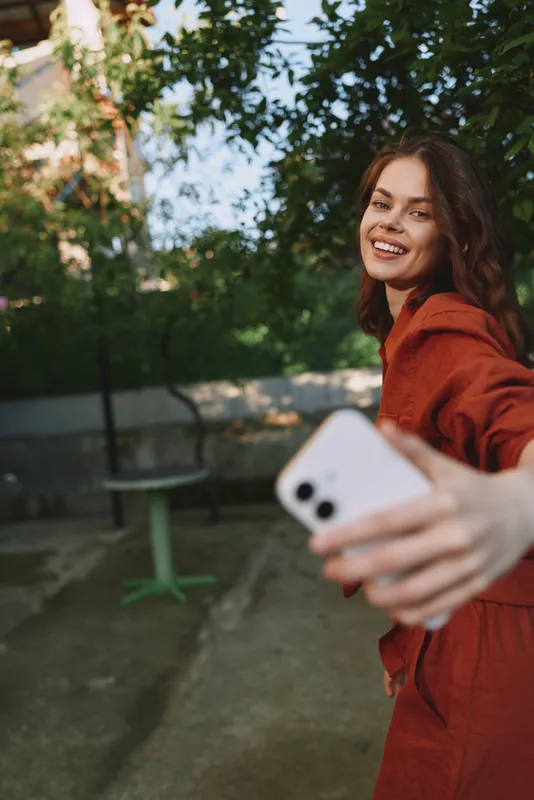 A smiling woman in a red outfit takes a selfie outdoors in a sunlit park, capturing a casual lifestyle moment with natural scenery and a cheerful mood for lifestyle photography