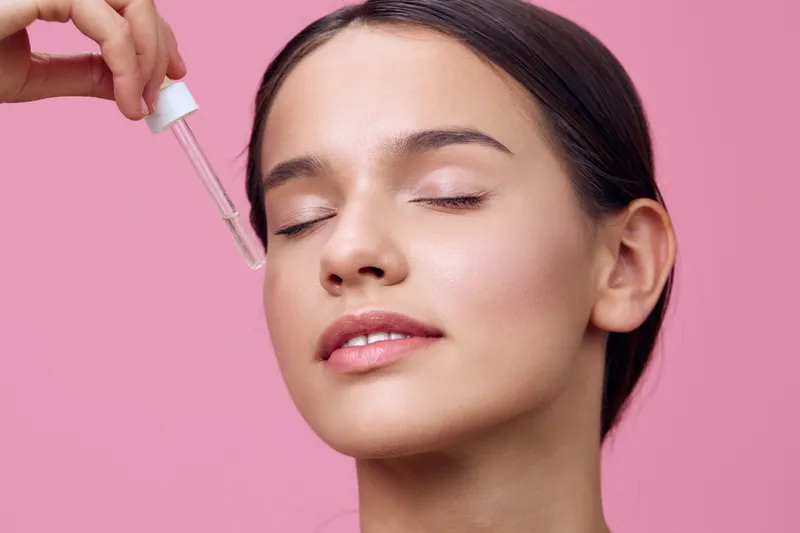 Young woman applying facial serum against a pink background, showcasing healthy skin and beauty routine, with a serene expression and natural cosmetics vibe